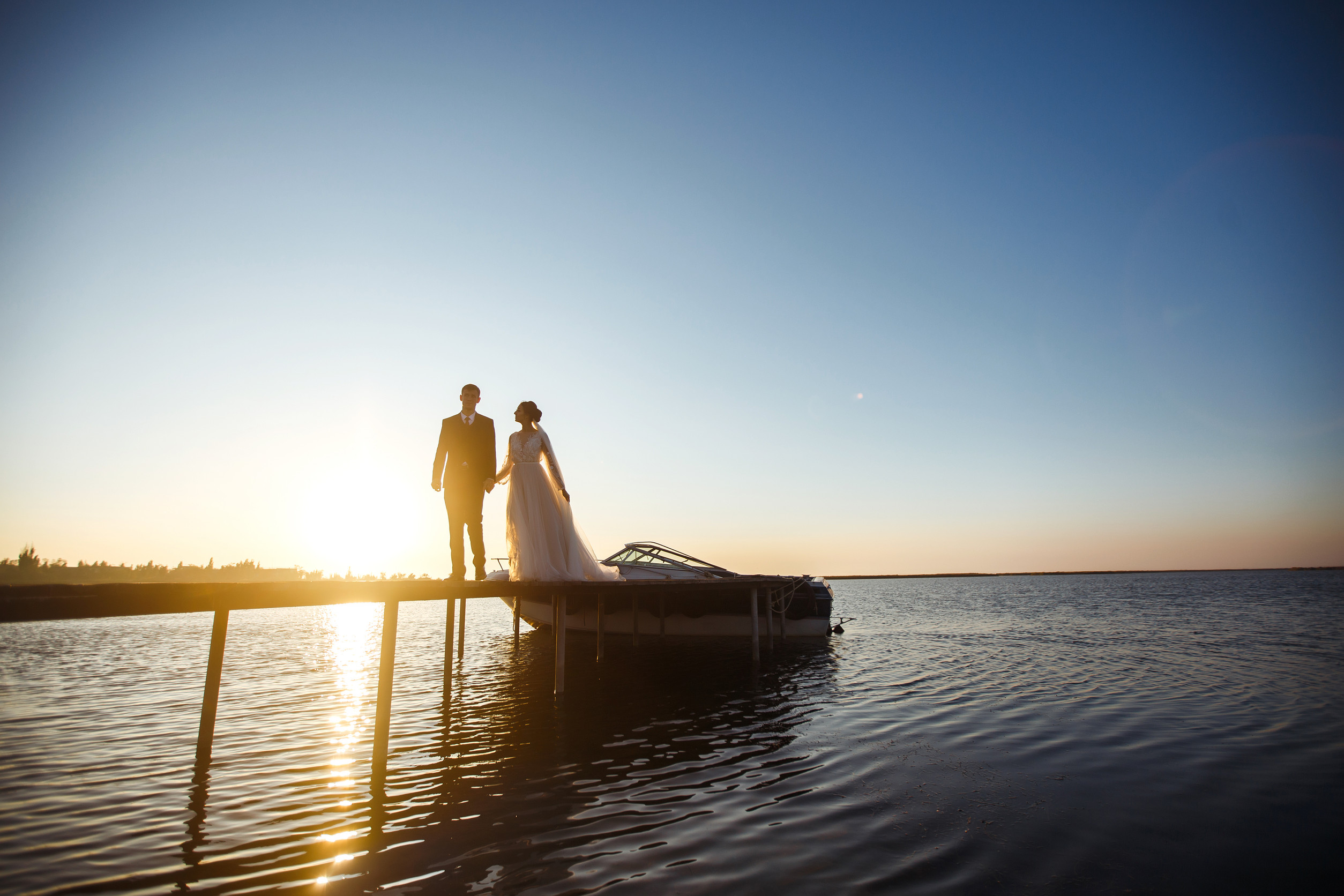 Beautiful bride and stylish groom together on the bridge against the background of the boat at sunset. Hochzeitsplanung Lakelove Wedding Hochzeitsplanerin für Hochzeiten